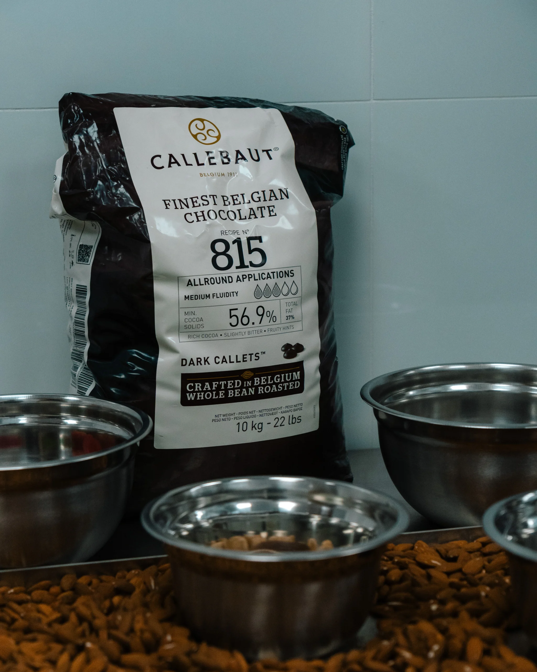 Επαγγελματική συσκευασία σοκολάτας Callebaut 815 δίπλα σε ανοξείδωτα μπολ και ξηρούς καρπούς σε πάγκο εργαστηρίου.
